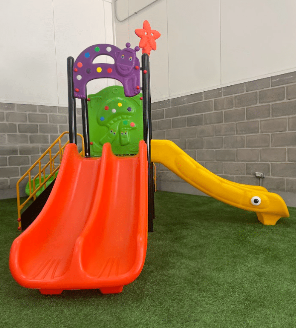 Estación de tobogán infantil colorida con tres toboganes en un patio interior con césped artificial.