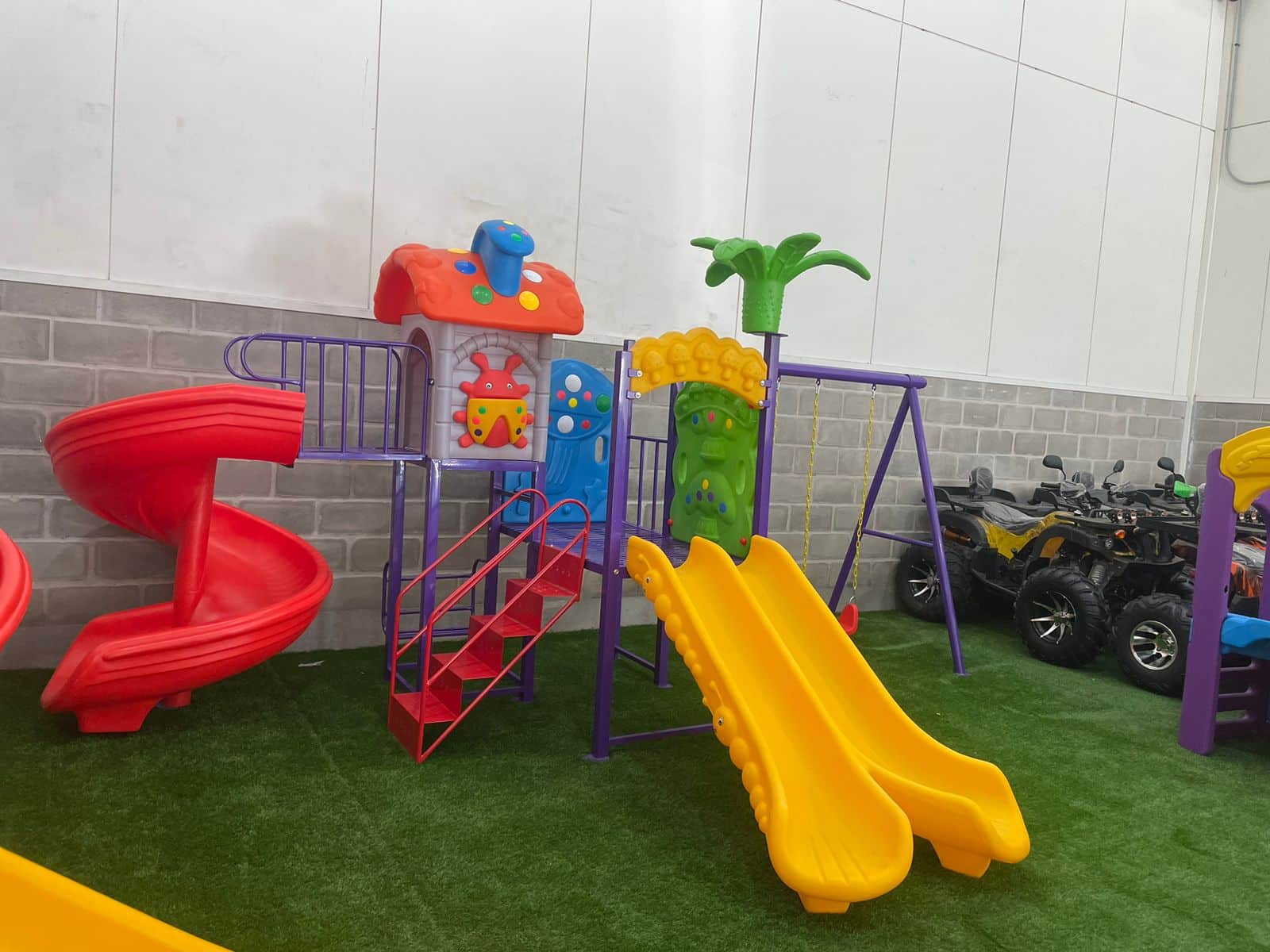 Juego infantil de exterior con toboganes rojos y amarillos, columpios y una casita de plástico colorida sobre césped artificial.